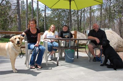 Four people and two dogs at a picnic table on the Mulligans patio, under a yellow umbrella with pines behind.