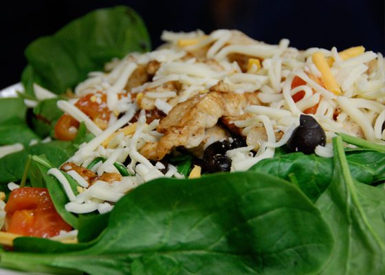A salad on a bed of spinach with grilled chicken, shredded cheese and black olives.