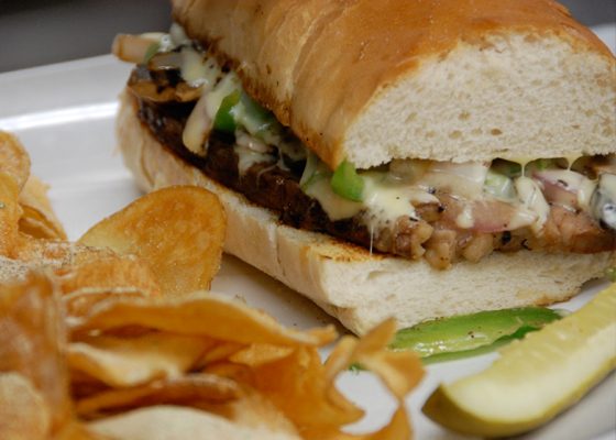 A steak sandwich on a long roll with peppers and onions, served with chippers and a pickle spear.