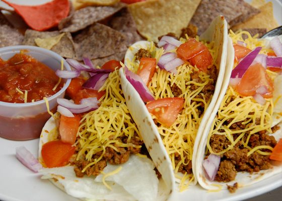 Two soft tacos loaded with shredded cheese, tomato and red onion, with tortilla chips and salsa.